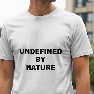 May include: White t-shirt with the text "UNDEFINED BY NATURE" in black. The t-shirt has short sleeves and a crew neck. The person wearing the shirt is standing outside in front of a building.
