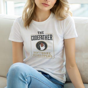Könnte beinhalten: Ein weißes kurzärmeliges T-Shirt mit einem Grafikdruck, der den Text THE CODEFATHER SOFTWARE DEVELOPERS in Schwarz und Gold zeigt. Das Design enthält ein kreisförmiges Emblem mit einer Hand, die eine Computermaus mit Kabel hält.