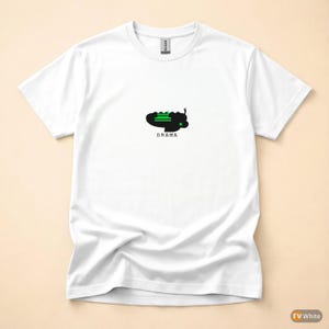 Könnte beinhalten: Weißes T-Shirt mit einem grafischen Design. Das Design zeigt eine schwarze Form mit grünen Linien und das Wort "DRAMA" in Schwarz. Das Shirt besteht aus einem weichen Material und hat einen Rundhalsausschnitt.