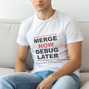 May include: White t-shirt with the text "MERGE NOW DEBUG LATER" in black and red, with "Teamwork In Progress" below. The shirt is worn with blue jeans.