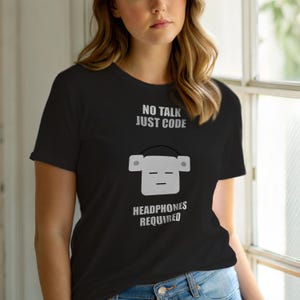 Op de afbeelding: Zwart T-shirt met de tekst "NO TALK JUST CODE" boven een grijze afbeelding van een koptelefoon. Daaronder staat "HEADPHONES REQUIRED". Het shirt heeft een ronde hals en korte mouwen.