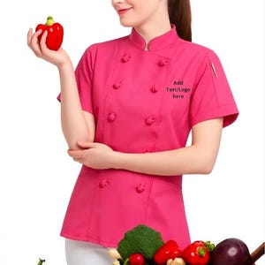 May include: A woman in a pink chef's jacket, holding a red pepper. The jacket has a stand-up collar and decorative buttons. The text "Add Text/Logo here" is on the jacket. A basket of vegetables is on a wooden surface.
