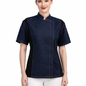 Puede incluir: Una chaqueta de chef de manga corta con un panel frontal de mezclilla azul oscuro con costuras naranjas en contraste, cuello mao y paneles laterales negros sólidos. La prenda incluye un pequeño bolsillo utilitario en la manga izquierda.