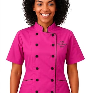 Pode incluir: Um casaco de chef rosa brilhante com acabamentos e botões pretos. O casaco de manga curta tem gola mandarim e design de abotoamento duplo. O texto "Add Text/Logo here" está bordado no lado direito. O casaco é usado por uma pessoa com cabelo encaracolado escuro.