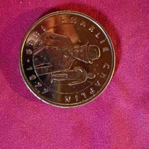 Puede incluir: Una moneda dorada que representa a Charlie Chaplin con su bombín y traje icónicos. La moneda está inscrita con "CHARLIE CHAPLIN" y los años "1889-1977". La moneda está sobre un fondo rosa.