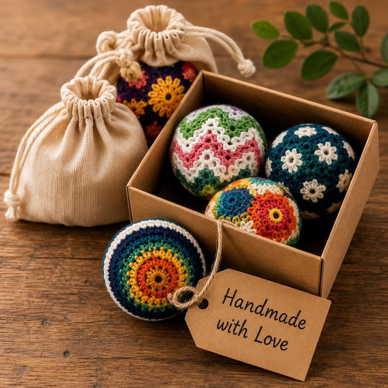 May include: A set of crocheted spheres in various colors and patterns, including floral and chevron designs, housed in a brown cardboard box and two beige drawstring pouches. A brown paper tag attached to the box reads Handmade with Love.