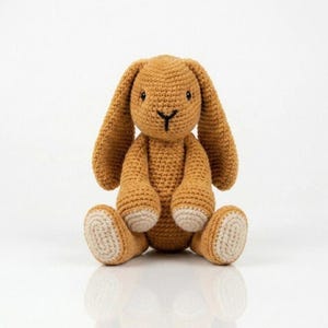 Könnte beinhalten: Ein handgehäkelter Hase in sitzender Position. Der Hase ist hauptsächlich in einem warmen Braunton gehalten, mit langen Ohren, einer schwarzen Nase und hellbeige Akzenten an den Füßen. Das Spielzeug ist aus weichem Garn gefertigt und für Kinder geeignet.
