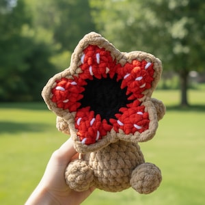 May include: A crocheted Demogorgon plushie. The plushie features a large, open, red and white flower-like head with a black center. The body is a light brown color, with two small, round appendages. The background is a blurred green.