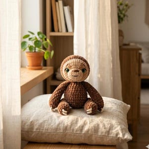 Op de afbeelding: Een gehaakt luiaard-speelgoed in bruin en beige, met groene ogen, zit op een beige kussen. De achtergrond toont een houten plank met boeken, een plant in een pot en een raam met transparante gordijnen.