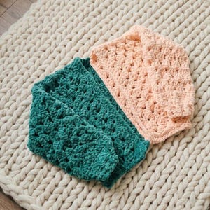 May include: Two crocheted sweaters in teal and peach colors are displayed on a cream-colored, chunky knit blanket. The sweaters have a textured, open stitch pattern. The teal sweater is folded, and the peach sweater is laid flat.