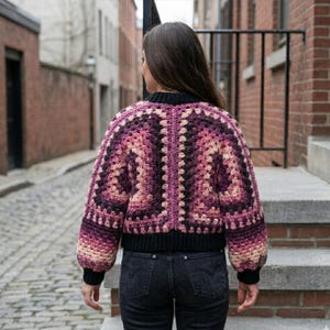 May include: A crocheted jacket in shades of purple, pink, and black. The jacket features a geometric pattern, a black collar, and black cuffs. The sleeves are long and slightly puffed. The person is wearing black jeans.