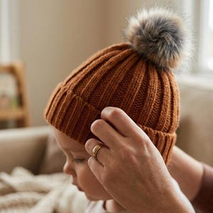Könnte beinhalten: Eine braune Strickmütze mit einem Kunstfell-Bommel. Die gerippte Strickmütze ist ein warmes, stilvolles Accessoire. Der Bommel ist eine Mischung aus Braun und Grau. Die Mütze ist ein Winteraccessoire.