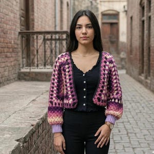 May include: A cropped, open-front crochet cardigan in shades of purple, pink, and cream. The sleeves have a gradient effect, transitioning from cream to purple. The cardigan is worn over a black top and paired with black pants.