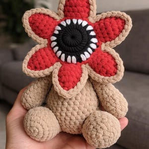 May include: A handmade crochet Demogorgon plush toy. The toy has a red, flower-shaped head with a black centre and white teeth. The body and limbs are tan, giving a soft, textured look.