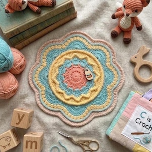 May include: A handmade, colorful crochet mat with a floral design, featuring shades of pink, yellow, and blue. The image also includes yarn balls, wooden blocks, a crochet fox, a wooden bunny, and a book that says "C is Croch".