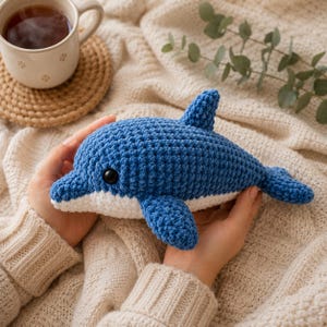 May include: A handmade crochet dolphin plush toy featuring a deep blue upper body and a white underbelly. The stuffed animal has a prominent dorsal fin, two side fins, a tail, and a single visible black plastic eye on the side of its head.