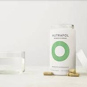 May include: A white cylindrical bottle of Nutrafol Women's Vegan supplements with a green circular design and the text "Nutrafol". Several brown capsules are next to the bottle and a clear glass of water.