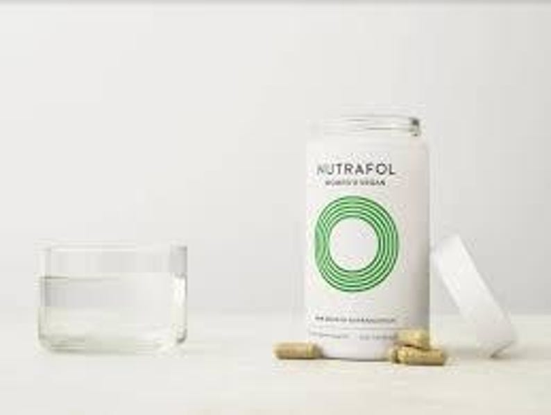 May include: A white cylindrical bottle of Nutrafol Women's Vegan supplements with a green circular design and the text "Nutrafol". Several brown capsules are next to the bottle and a clear glass of water.