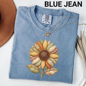Puede incluir: Una camiseta azul vaquero con un diseño de girasol. El girasol tiene un diseño de patchwork con varios patrones y colores, incluyendo naranja, amarillo y verde. La camiseta tiene cuello redondo y mangas cortas. El texto "BLUE JEAN" está en la parte superior de la imagen.