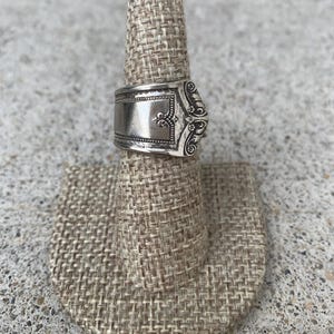 May include: A silver ring with an ornate design, featuring a rectangular face with detailed patterns and a decorative edge. The ring is displayed on a beige ring holder, showcasing its intricate craftsmanship and vintage style.