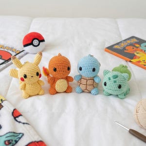 May include: Four crocheted Pokémon characters: Pikachu, Charmander, Squirtle, and Bulbasaur. Each character is a different color, with black eyes and small details. A Poké Ball, a Pokémon book, and a blanket are also visible.