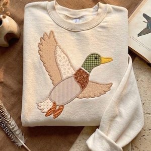 May include: A cream-colored crewneck sweatshirt featuring a large, central appliqué of a flying mallard duck. The duck is constructed from various fabric patches in shades of brown, tan, and green, showcasing a mix of floral, striped, and checkered patterns.