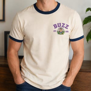 May include: A cream-colored ringer t-shirt with dark blue trim on the collar and sleeves. The chest features a graphic of the character Buzz Lightyear with the purple text BUZZ above and the text EST. 1995 on either side of the character.