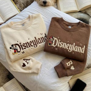 May include: Two crewneck sweatshirts, one cream and one brown, each featuring embroidered Disneyland text, small star and butterfly accents, and a character. The cream sweatshirt shows Mickey Mouse and the letter A, while the brown one shows Minnie Mouse and the letter B.