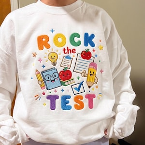 以下が含まれることがあります： カラフルな刺繍デザインが施された白いスウェットシャツ。「ROCK the TEST」の文字と、電球、リンゴ、鉛筆、本の画像が含まれています。スウェットシャツはクルーネックです。
