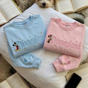 May include: A light blue sweatshirt and a light pink sweatshirt, each featuring embroidered Disneyland text, small stars, and butterflies. The blue sweatshirt displays Mickey Mouse, while the pink one shows Minnie Mouse. Each sleeve has a letter and heart.