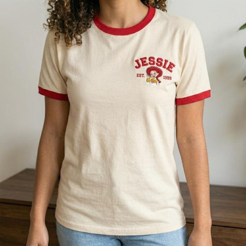May include: A cream-colored ringer t-shirt with red trim on the collar and sleeve cuffs. The chest features a graphic of the character Jessie with the text "JESSIE" in red arched lettering above and "EST. 1999" printed in smaller red text below.