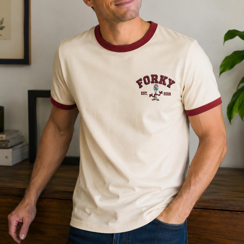 May include: A cream-colored ringer t-shirt with dark red trim on the collar and sleeve cuffs. The chest features a small graphic with the text "FORKY" in a collegiate font, "EST. 2019" below it, and a small illustration of a spork character in between.