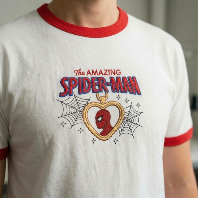 Op de afbeelding: Een wit ringer-t-shirt met rode boorden bij de kraag en mouwen. Op de borst staat een geborduurd ontwerp met de tekst The AMAZING SPIDER-MAN in rood en blauw, een gouden hartvormige hanger met een rode Spider-Man-kop en spinnenwebben.