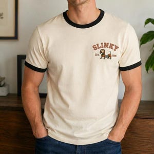 May include: A cream-colored ringer t-shirt with black trim on the crew neckline and short sleeves. The chest features a graphic of a brown dachshund dog with the text SLINKY arched above it and the text EST. 1995 positioned below the dog illustration.