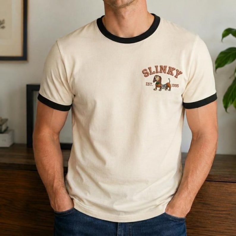 May include: A cream-colored ringer t-shirt with black trim on the crew neckline and short sleeves. The chest features a graphic of a brown dachshund dog with the text SLINKY arched above it and the text EST. 1995 positioned below the dog illustration.