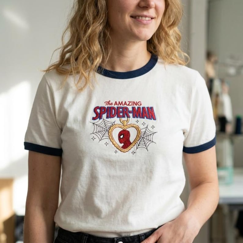 Op de afbeelding: Een wit ringer-t-shirt met marineblauwe biesjes bij de kraag en mouwen. Op de borst staat een geborduurd ontwerp van een gouden hartvormige hanger met een rood Spider-Man-masker, geflankeerd door spinnenwebben en de tekst The AMAZING SPIDER-MAN.
