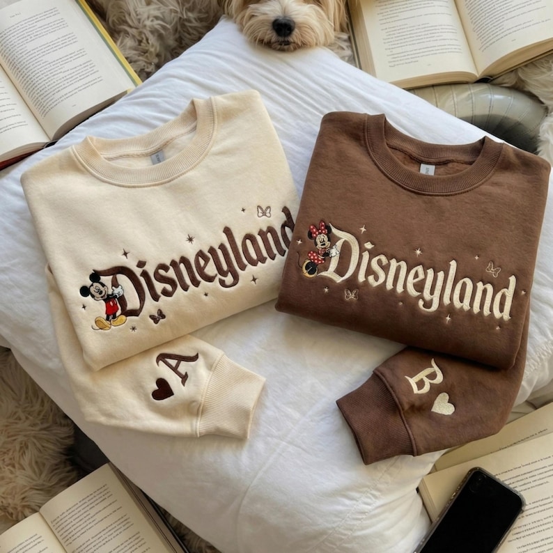 May include: Two crewneck sweatshirts, one cream and one brown, each featuring embroidered Disneyland text, small star and butterfly accents, and a character. The cream sweatshirt shows Mickey Mouse and the letter A, while the brown one shows Minnie Mouse and the letter B.