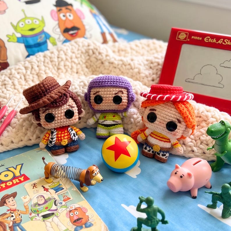Może przedstawiać: Szydełkowe postacie z Toy Story, w tym Woody, Buzz Lightyear i Jessie, wraz z piłką, zabawką jamnikiem, świnką skarbonką i zielonymi żołnierzykami. Zabawki są ułożone na niebieskiej powierzchni z książką Toy Story i czerwoną zabawką Etch-A-Sketch.