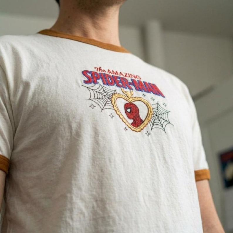 Op de afbeelding: Een wit ringer-t-shirt met bruine boorden voorzien van een geborduurd ontwerp met de tekst The Amazing Spider-Man boven een goudkleurig hartvormig medaillon met een rode Spider-Man-figuur, geflankeerd door zwarte spinnenwebben en sterretjes.