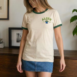 May include: A cream-colored ringer t-shirt with dark green trim on the collar and sleeves. The chest features a graphic with the text ALIEN, a small green three-eyed character, and the text EST. 1995. The shirt is paired with a blue denim skirt.
