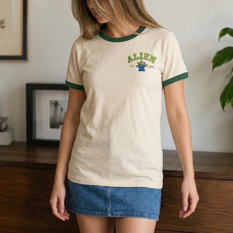 May include: A cream-colored ringer t-shirt with dark green trim on the collar and sleeves. The chest features a graphic with the text ALIEN, a small green three-eyed character, and the text EST. 1995. The shirt is paired with a blue denim skirt.