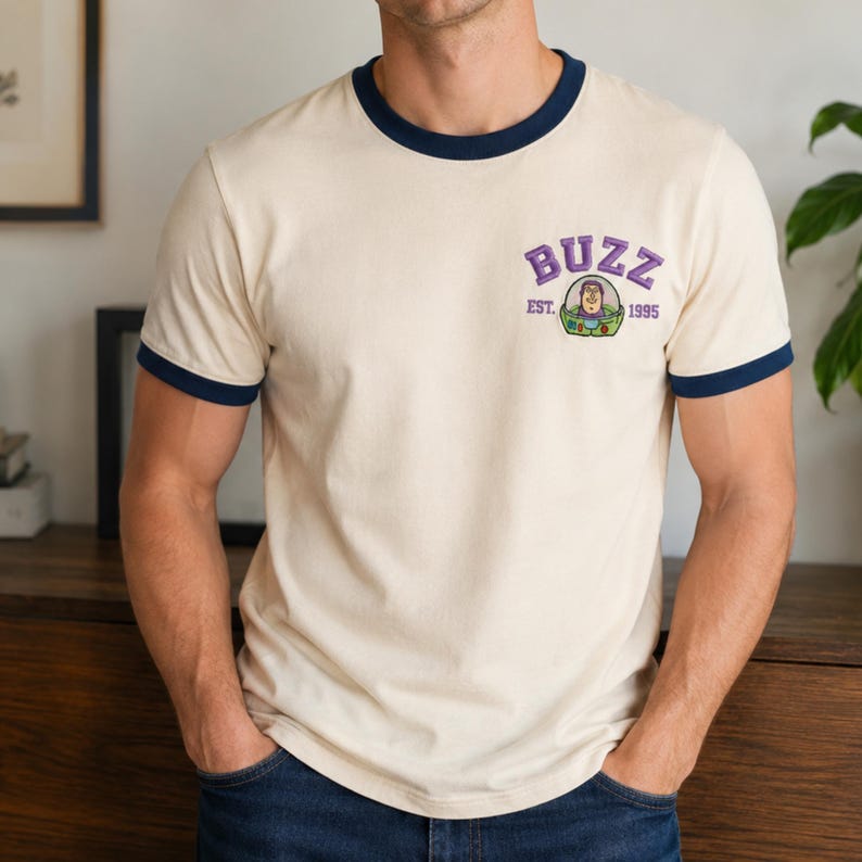 May include: A cream-colored ringer t-shirt with dark blue trim on the collar and sleeves. The chest features a graphic of the character Buzz Lightyear with the purple text BUZZ above and the text EST. 1995 on either side of the character.