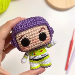 Może przedstawiać: Ręcznie robiona szydełkowa lalka Buzz Lightyear. Lalka ma fioletowy hełm, czarne oczy oraz zielono-biały kombinezon kosmiczny. Lalka jest trzymana w dłoni, co pokazuje szczeg&oacute;łowe rzemiosło i żywe kolory.