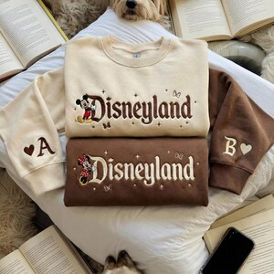 May include: Two crewneck sweatshirts featuring embroidered Disneyland text. The top sweatshirt is cream with brown text and a Mickey Mouse graphic. The bottom sweatshirt is brown with cream text and a Minnie Mouse graphic. Both have sleeve embroidery.