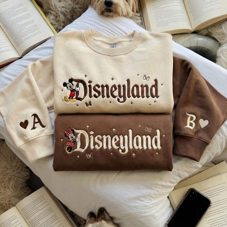 May include: Two crewneck sweatshirts featuring embroidered Disneyland text. The top sweatshirt is cream with brown text and a Mickey Mouse graphic. The bottom sweatshirt is brown with cream text and a Minnie Mouse graphic. Both have sleeve embroidery.