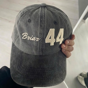 May include: A gray baseball cap with the word "Briar" embroidered in white script and the number "44" in a cream-colored block font. The cap has a curved brim and a button on top.