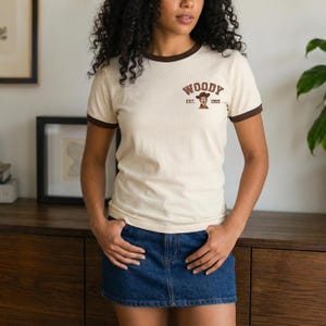 May include: A cream-colored ringer t-shirt with brown trim on the collar and sleeves. The chest features a graphic of a cowboy character with the text "WOODY" arched above and "EST. 1995" printed below the character in a brown, collegiate-style font.