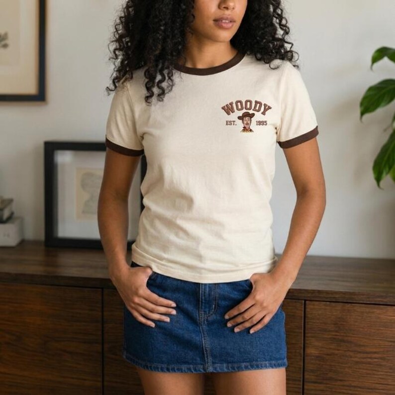 May include: A cream-colored ringer t-shirt with brown trim on the collar and sleeves. The chest features a graphic of a cowboy character with the text "WOODY" arched above and "EST. 1995" printed below the character in a brown, collegiate-style font.