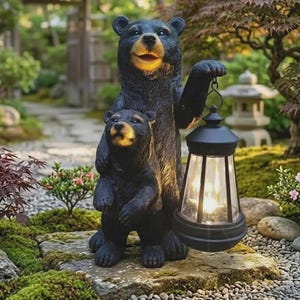 May include: A black bear statue featuring a large adult bear standing behind a smaller cub. The adult bear holds a black lantern with a glowing light in its raised paw. Both bears have textured fur, light-colored muzzles, and dark eyes.