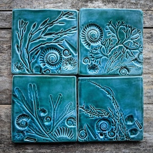 Puede incluir: Cuatro baldosas de cerámica turquesa, cada una con un diseño en relieve diferente de vida marina, incluyendo conchas en espiral, algas y coral. Las baldosas están dispuestas sobre una superficie de madera envejecida, creando un tema costero.